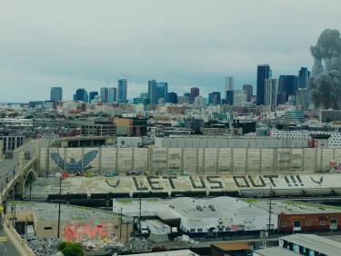 Los Angeles with many buildings, two columns of black smoke rising, and the words „LET US OUT“ painted on a wall